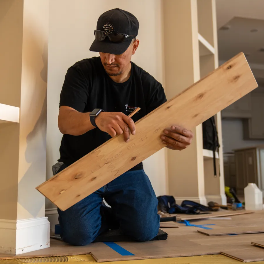 Man in black shirt and cap measuring wooden plank while installing flooring in home interior.