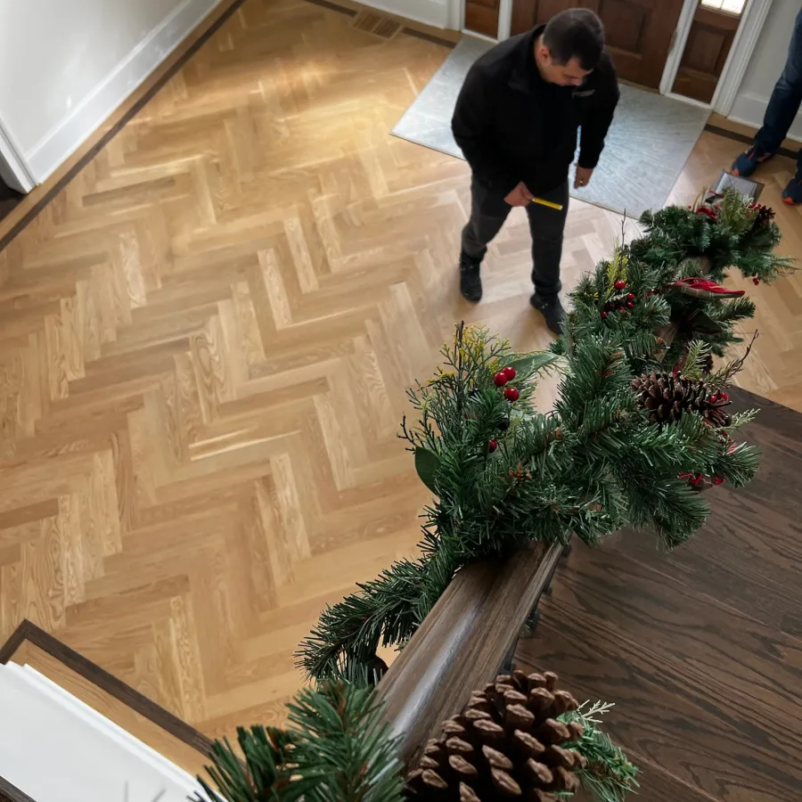 Man standing in a home foyer with wooden herringbone flooring and a staircase decorated with pine garland and pinecones.