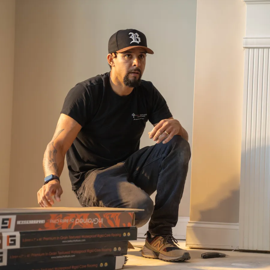 Man wearing a black cap and t-shirt crouching beside stacked flooring planks near a fireplace in a home.