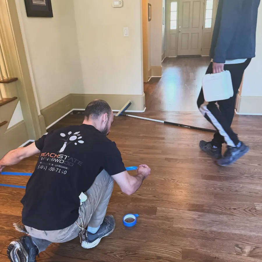 Worker taping hardwood floor inside a house hallway while another person walks past holding a toolbox.