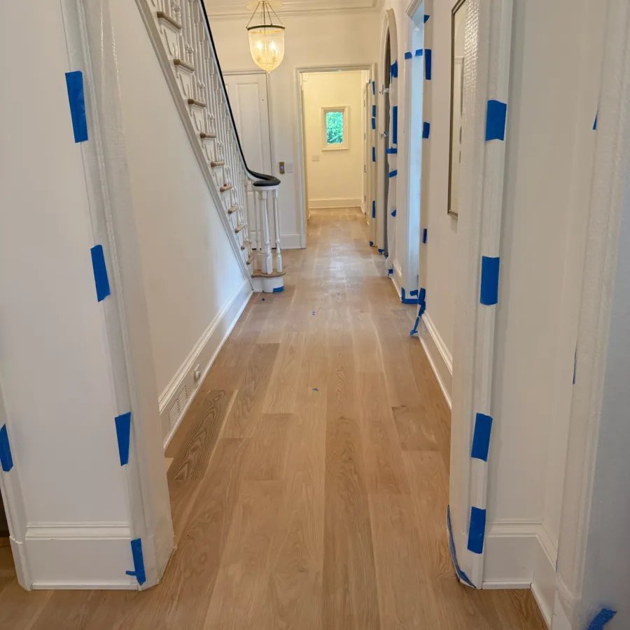 Long hallway with light wood flooring, white walls, staircase, and protective tape on door frames during renovation.