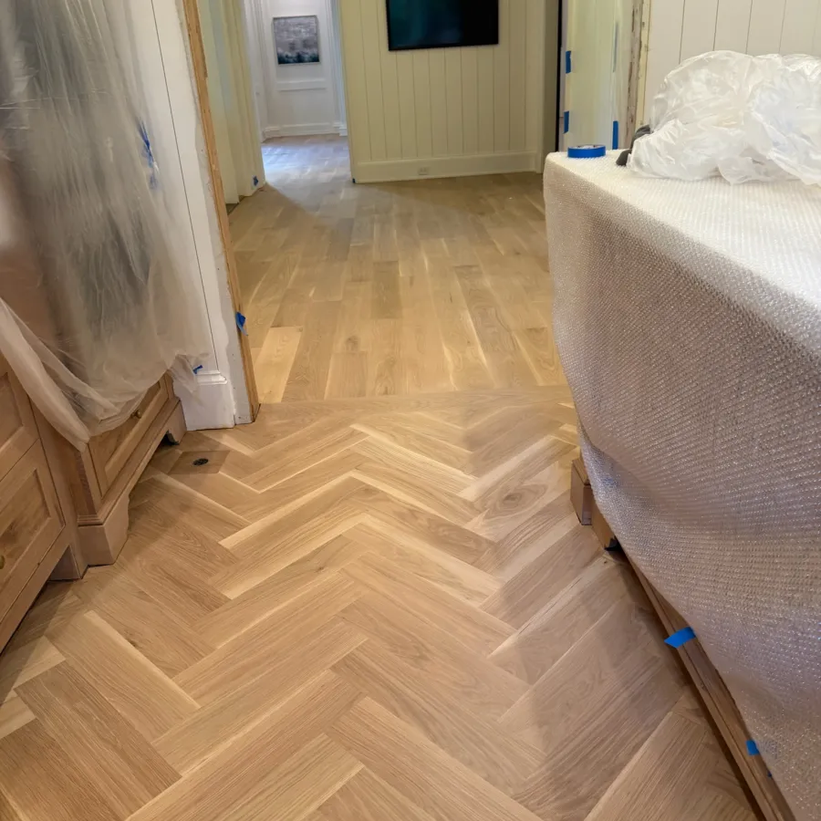 Light wood herringbone flooring transitions to plank flooring in a bright room with covered furniture and wall-mounted TV.