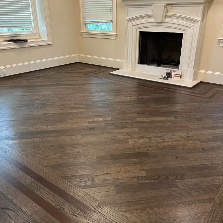 Empty room with dark patterned hardwood floor, white fireplace, beige walls, and two windows with blinds.