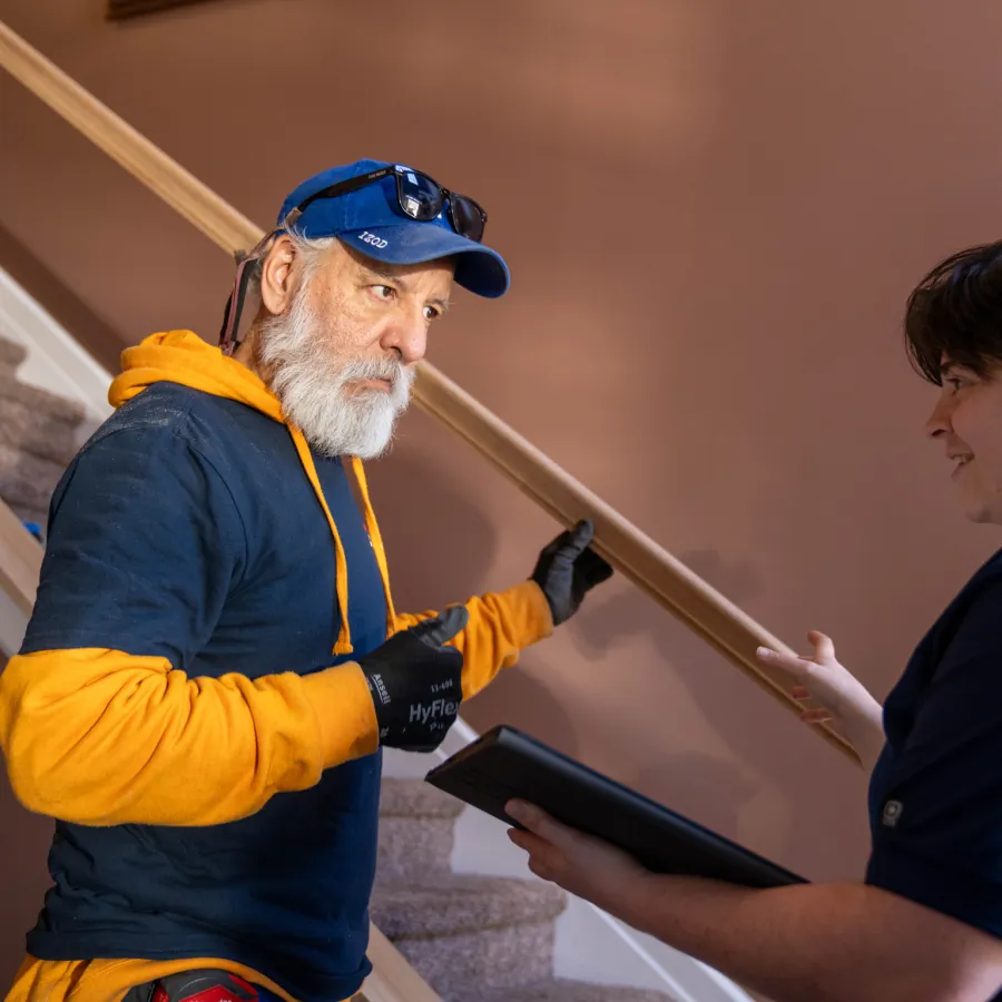 Older man in work gloves and cap discussing staircase project with younger person holding a tablet indoors