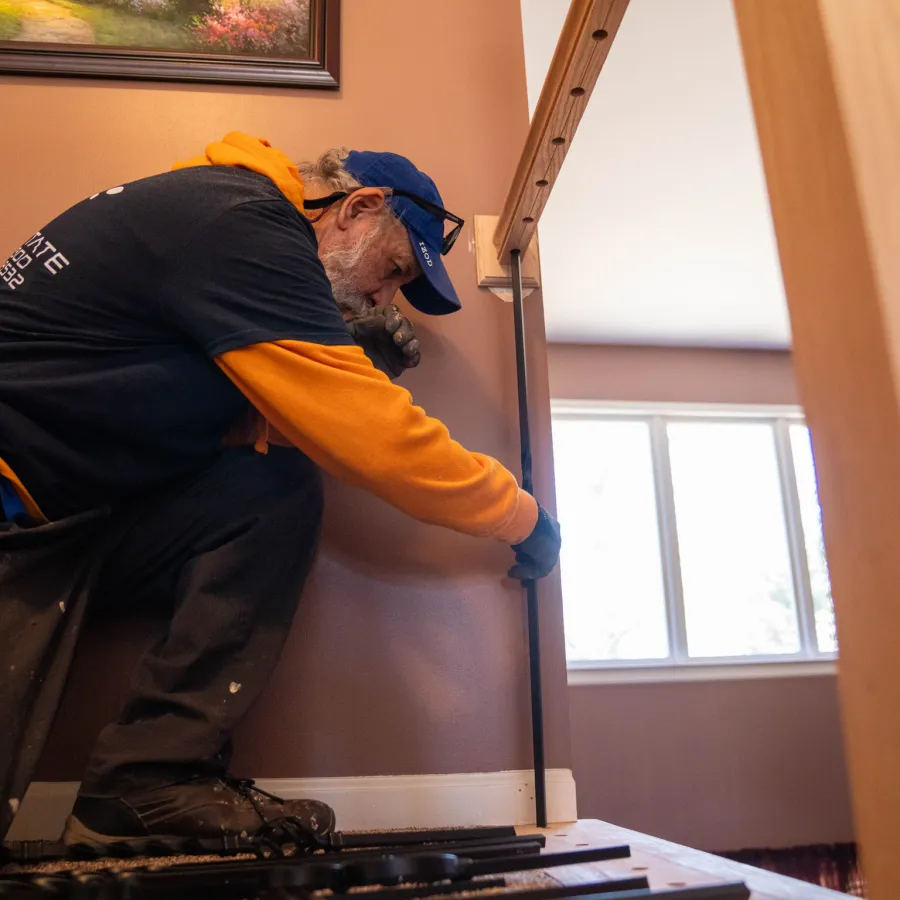 Older man in orange and black clothing installing a wooden handrail on an indoor staircase near a window.