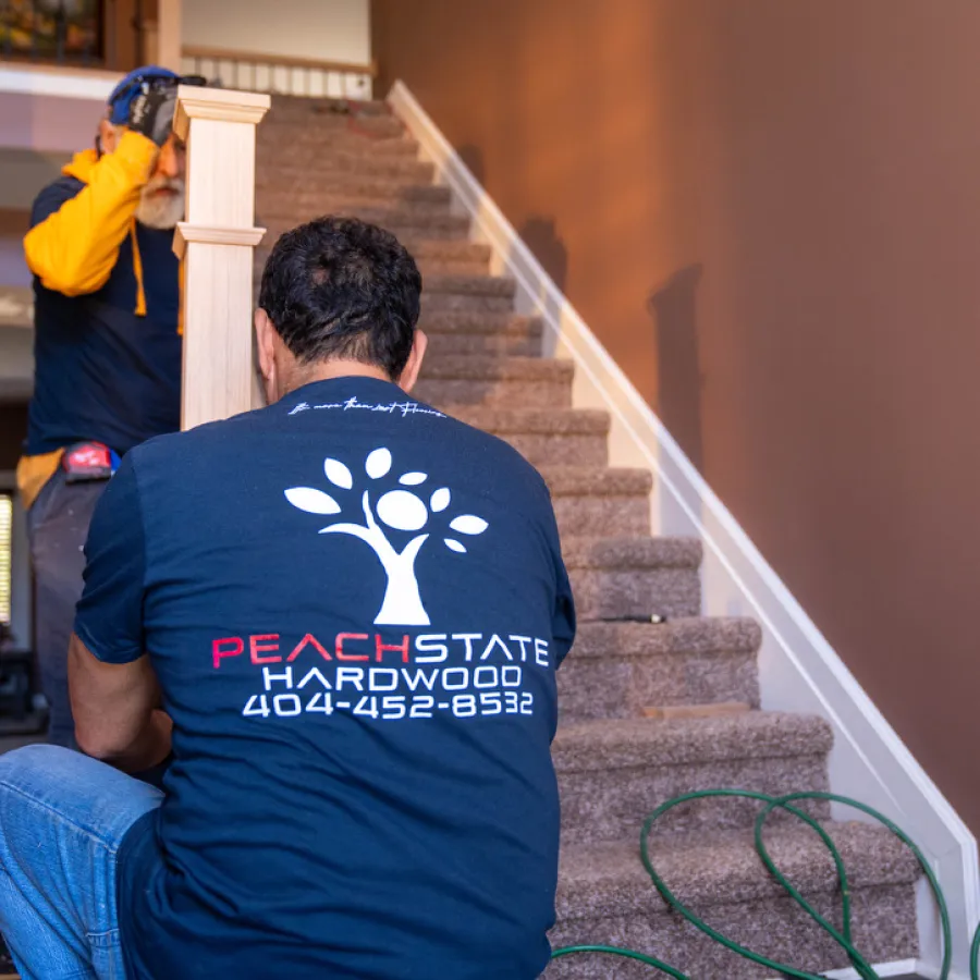 Carpenters installing hardwood flooring and staircase railing in a home interior with Peach State Hardwood branding.