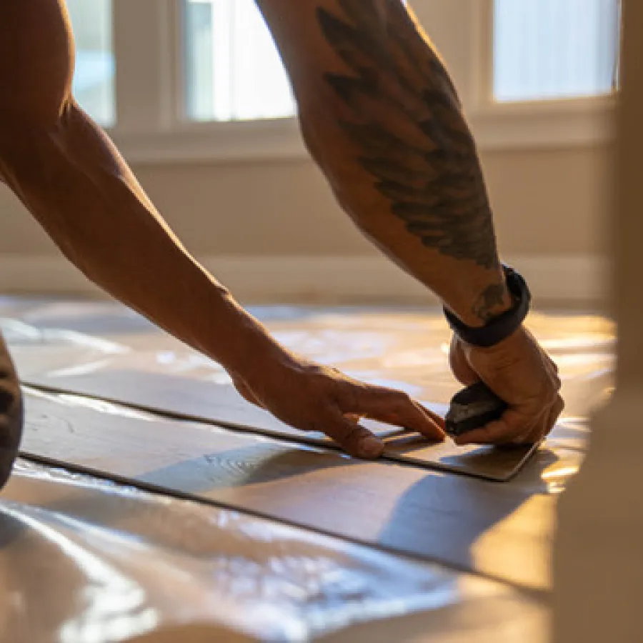Person installing laminate flooring, cutting pieces with a tool near a window in natural light.