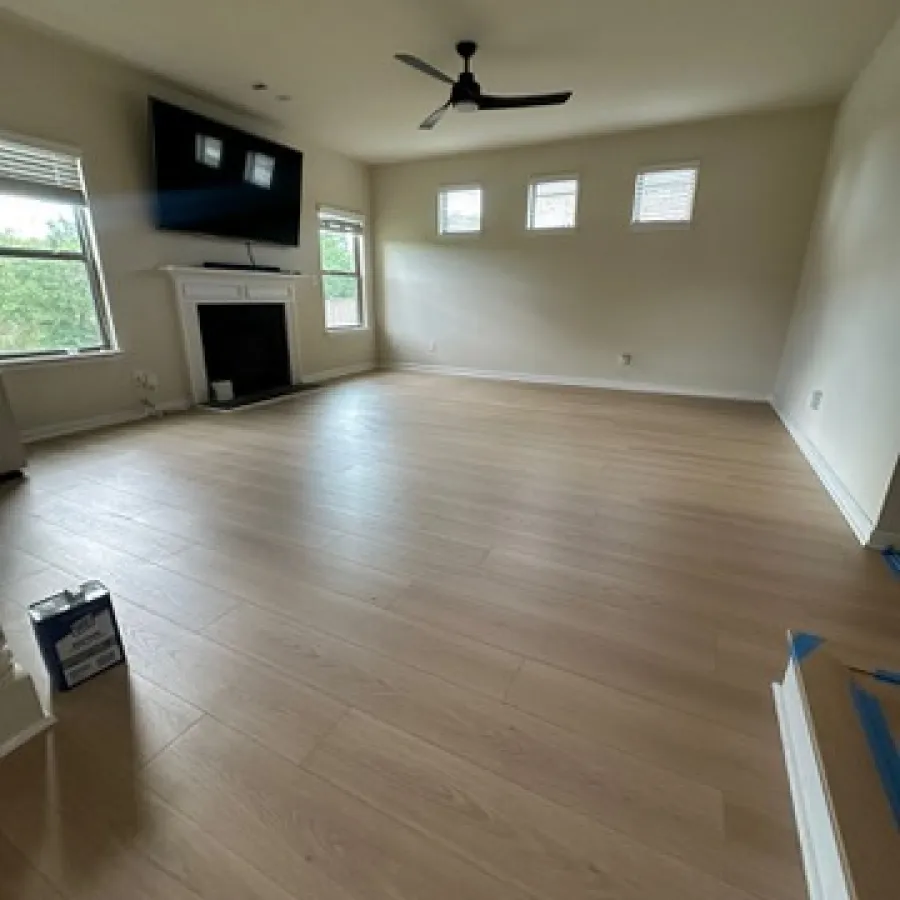 Empty living room with light wood flooring, ceiling fan, mounted TV, fireplace, and natural light from windows.