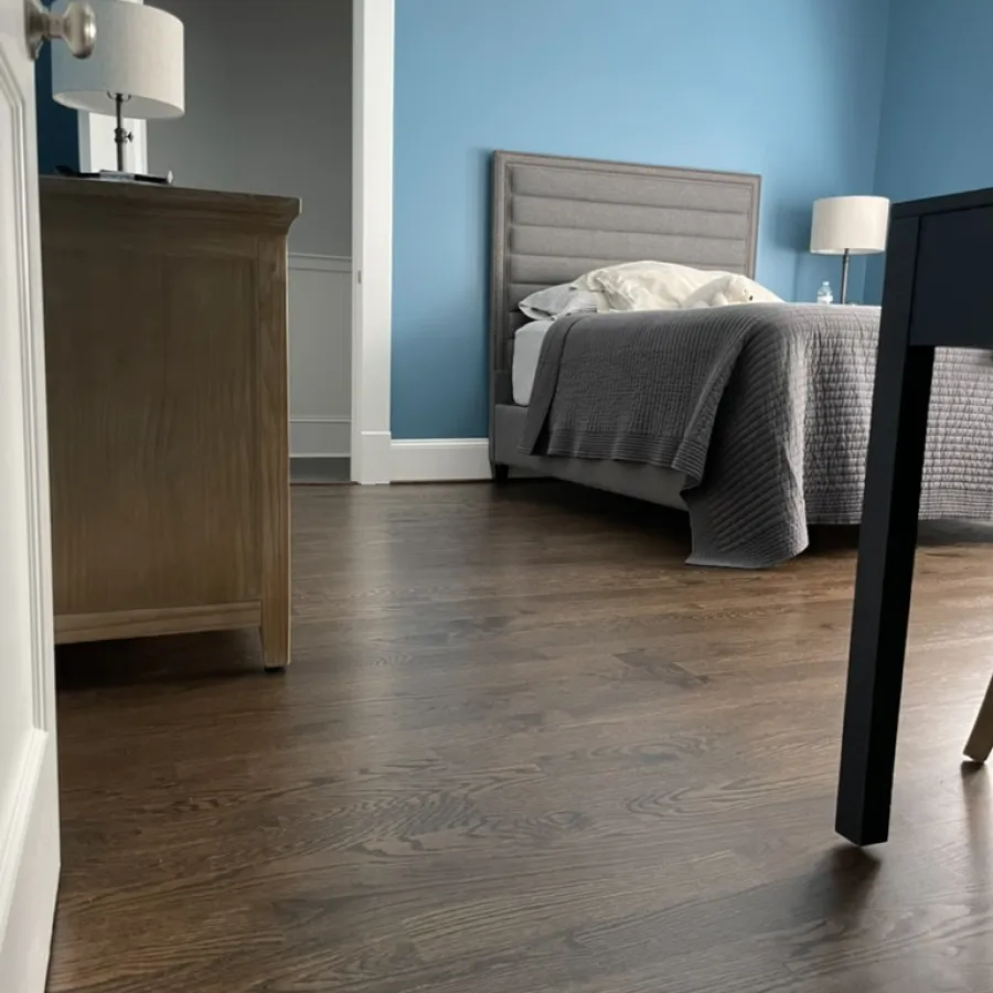 Modern bedroom with a gray upholstered bed, blue walls, wooden dresser, two lamps, and dark hardwood flooring.