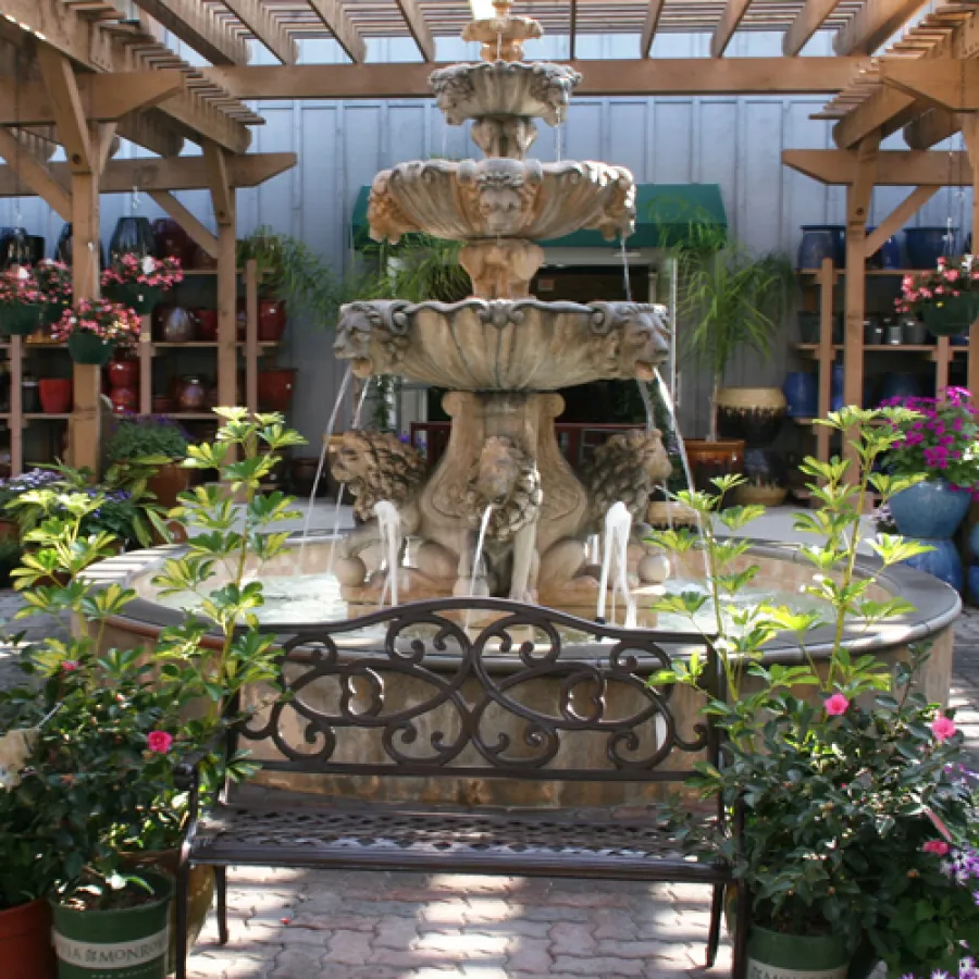 Ornate stone fountain with flowing water surrounded by potted plants and a wrought iron bench under wooden pergola.