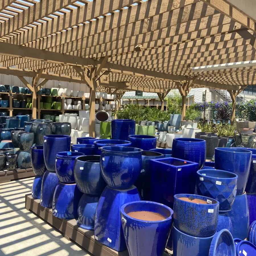 Outdoor display of various blue ceramic planters under wooden lattice shade structure in a garden center.