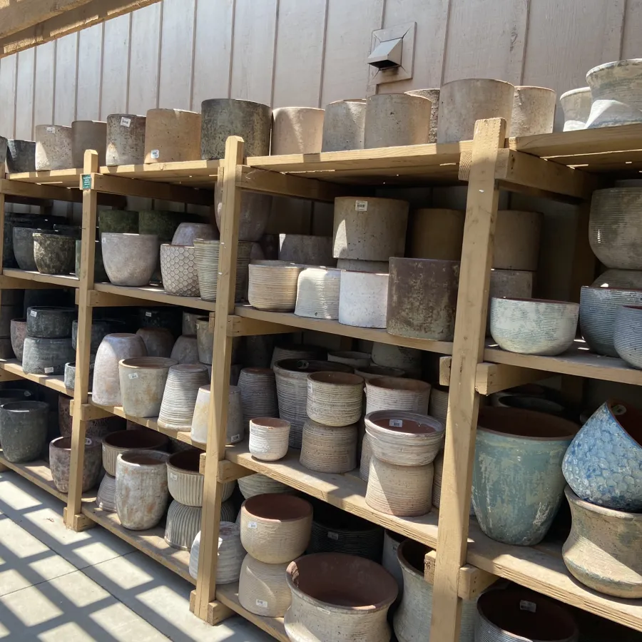 Various ceramic and stone plant pots displayed on wooden shelves in an outdoor garden center under sunlight.