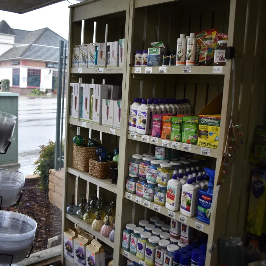 Shop shelves stocked with gardening supplies, fertilizers, plant care products, and decorative garden items near a rainy storefront.