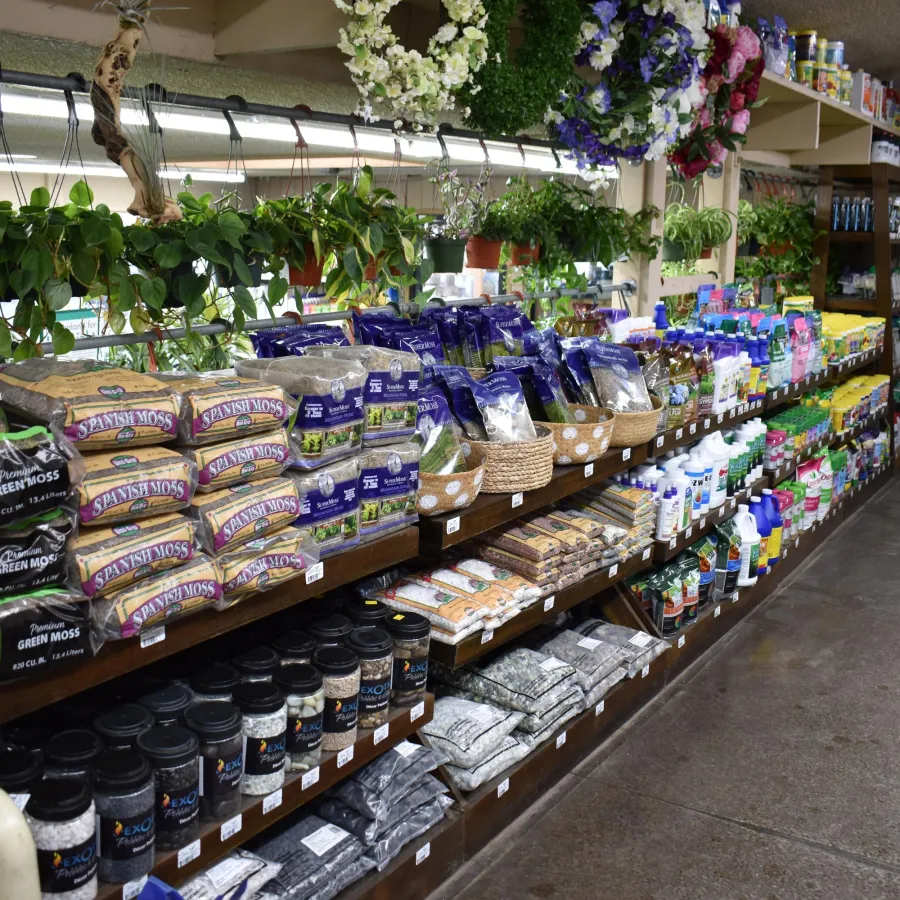 Store aisle with shelves stocked with gardening supplies, moss bags, fertilizers, and hanging plants under bright lights.