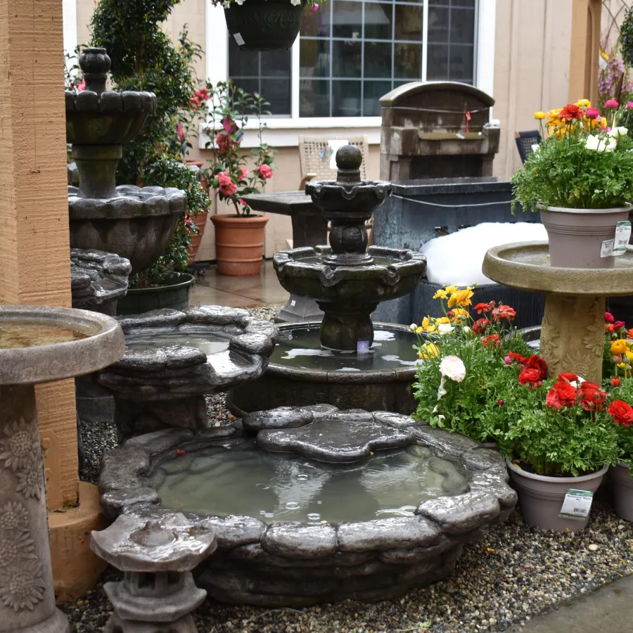 Garden center display featuring stone fountains and colorful potted flowers on gravel ground near a building.