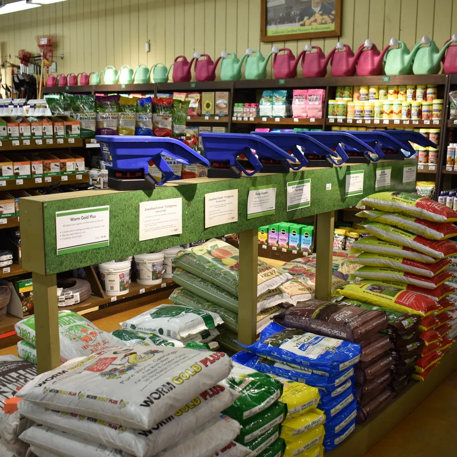 Garden center aisle with soil, fertilizer bags, watering cans, and gardening tools on shelves.