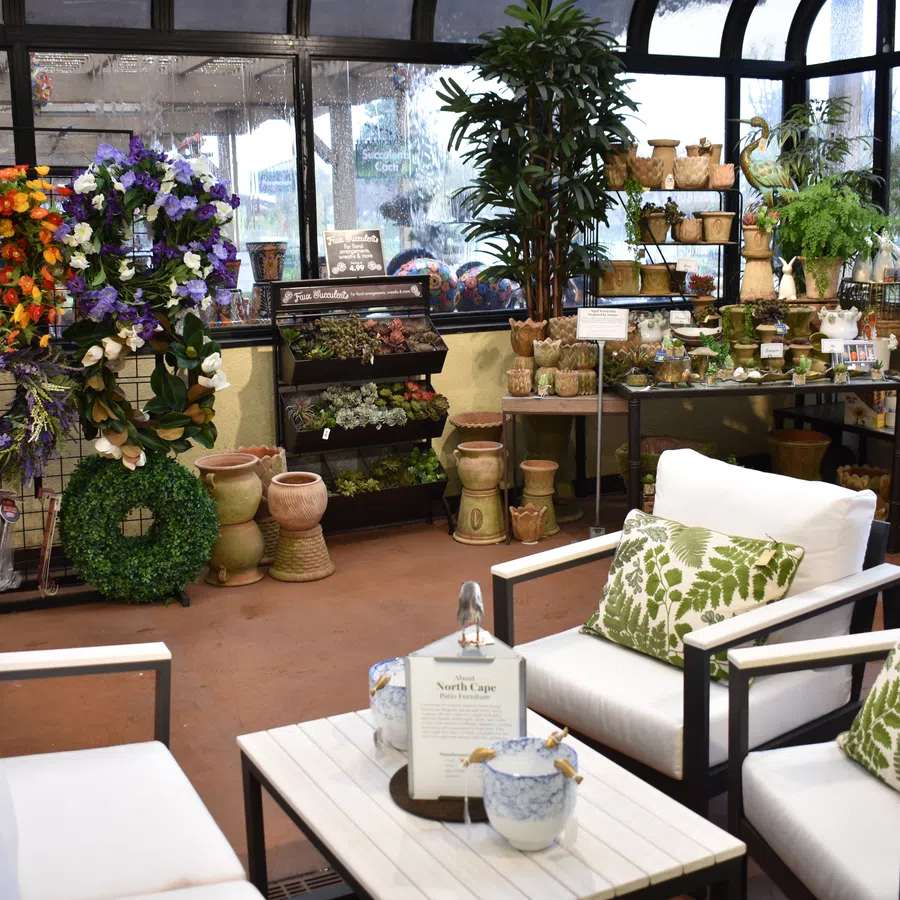 Cozy indoor garden shop with white armchairs, leafy cushions, floral wreaths, potted plants, and pottery displays.