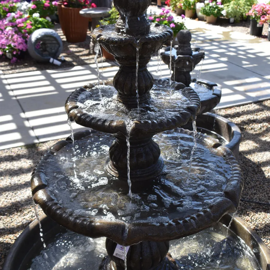 Three-tiered decorative water fountain in a sunny garden center surrounded by colorful flowers and plants.