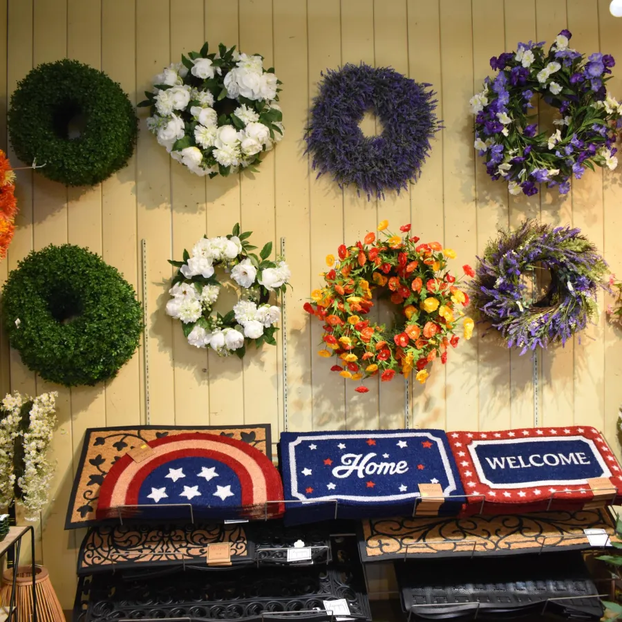 Colorful wreaths and patriotic-themed welcome mats displayed on a wall and shelf in a store.