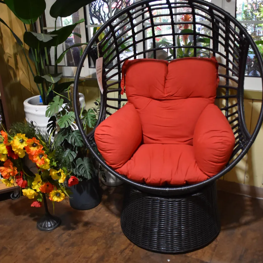 Black rattan round chair with bright red cushions next to potted plants and floral wreath indoors.