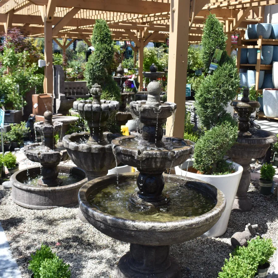 Outdoor garden center display with multiple stone water fountains, green plants, and wooden pergola overhead.