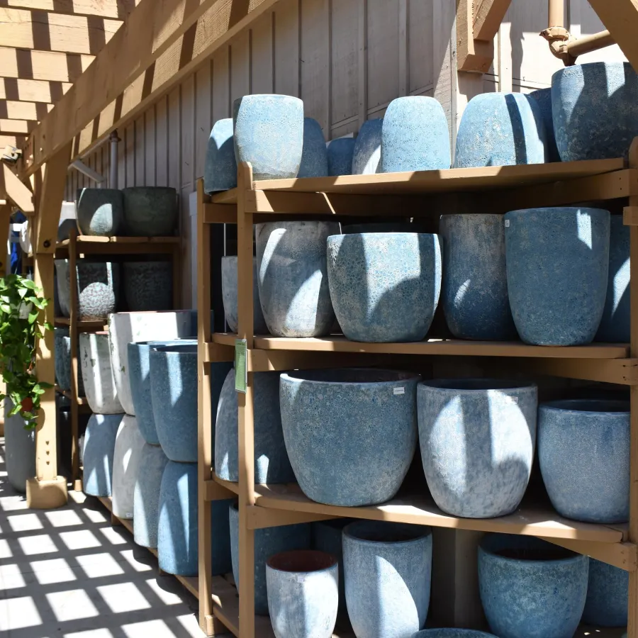 Shelves of various blue ceramic pots displayed under wooden pergola casting grid shadows.