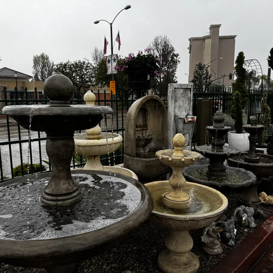 Variety of outdoor stone and concrete water fountains displayed in a rainy garden center with wet pavement and overcast sky