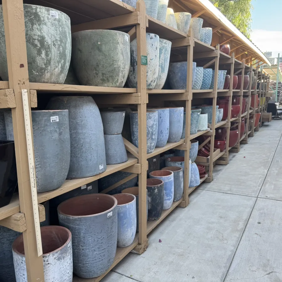 Rows of ceramic and stone plant pots displayed on wooden shelves in an outdoor garden center aisle.