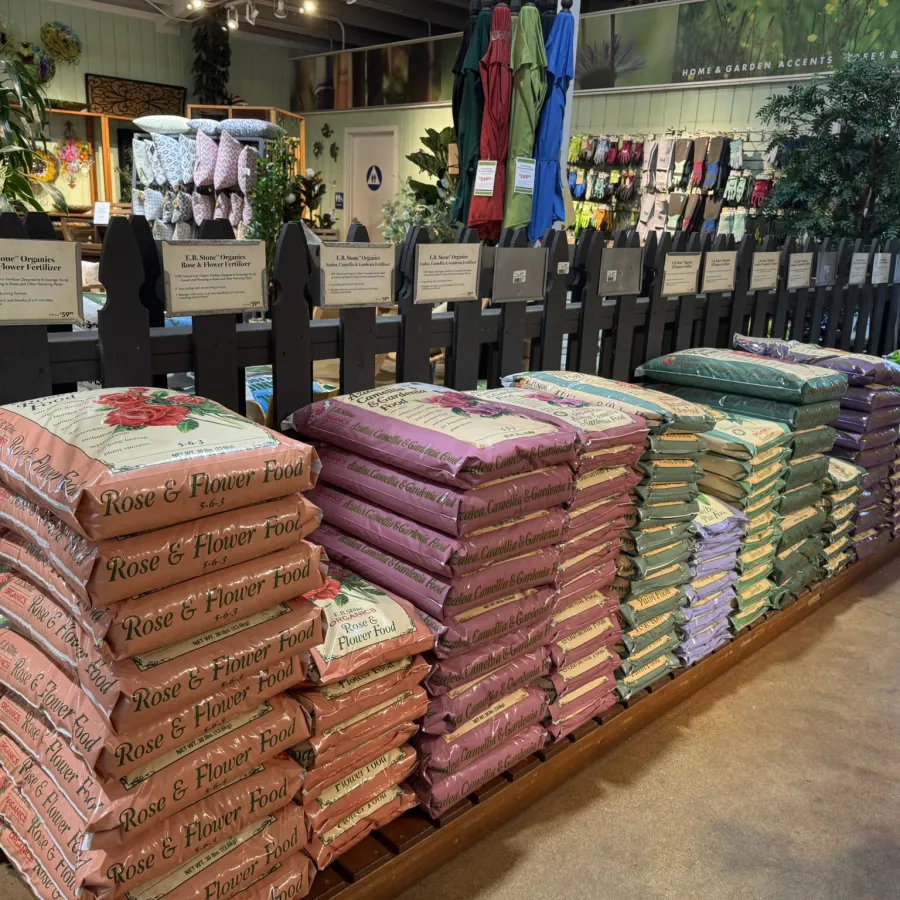 Stacks of colorful bags of rose and flower food fertilizer displayed on a wooden shelf in a garden store.