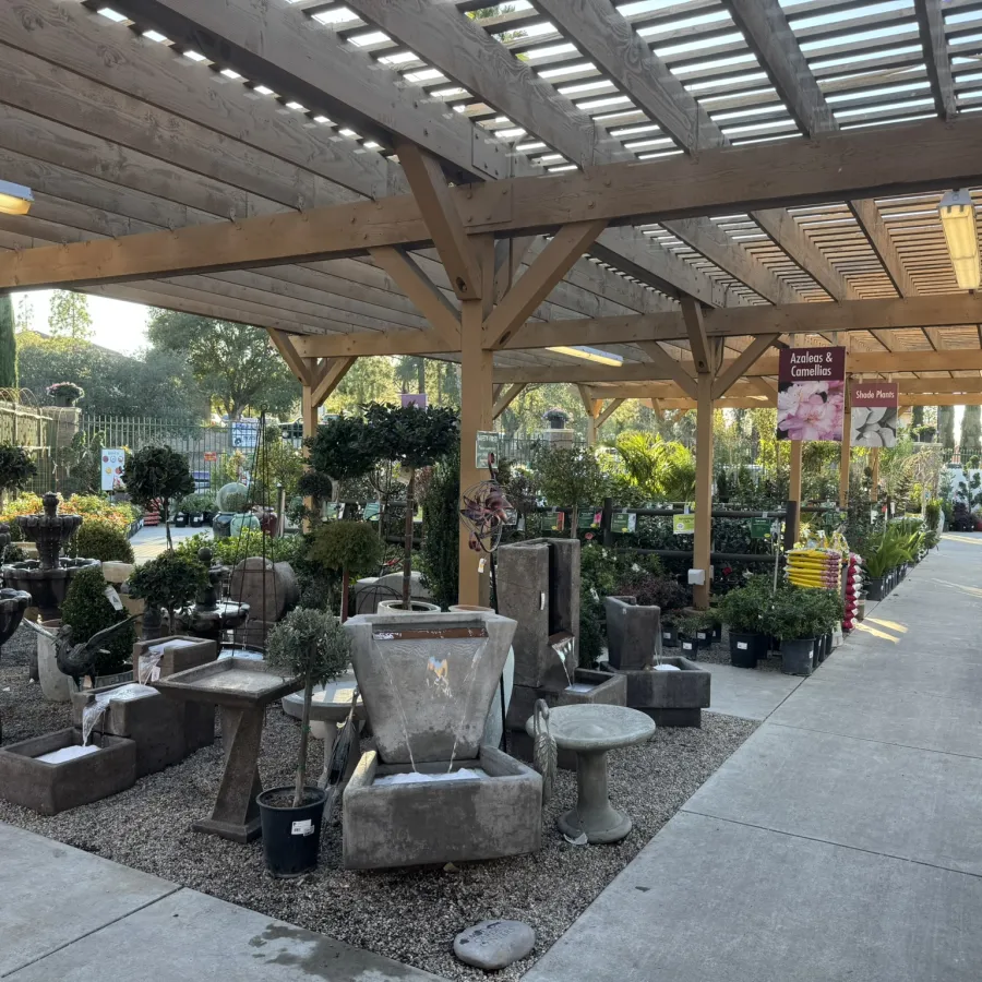Outdoor garden center with wooden pergola displaying fountains, plants, and garden decor along concrete pathways.