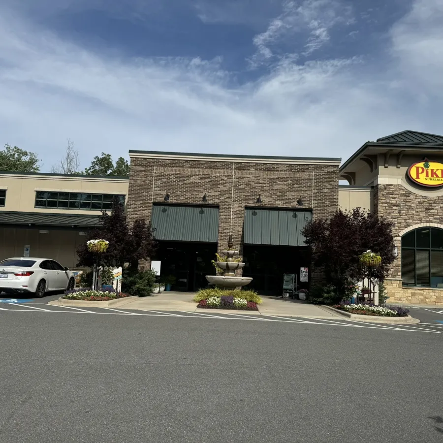 Brick and stone building with Pike Nurseries sign, fountain, and parked cars under blue sky with scattered clouds.