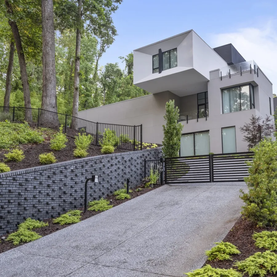 Modern multi-story home with cantilevered upper floor, landscaped yard, and sloped driveway with black fence gate.