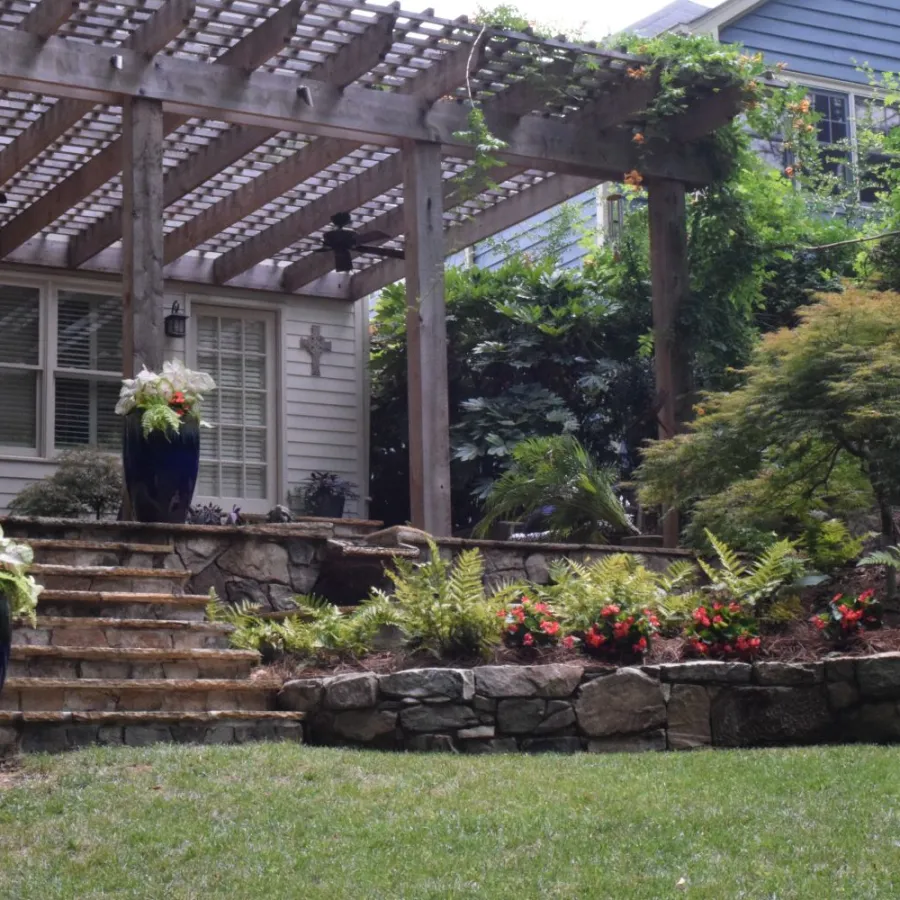 Stone terrace with flower pots, wooden pergola, stone steps, and lush garden in a residential backyard.