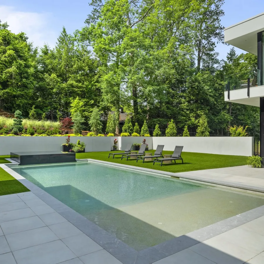Modern backyard with rectangular swimming pool, lounge chairs, green lawn, and white patio next to a house.