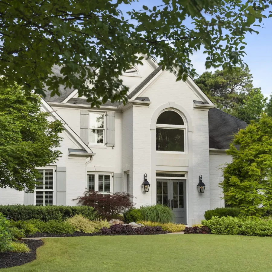 White two-story house with arched window, landscaped garden, and lush green lawn under blue sky.