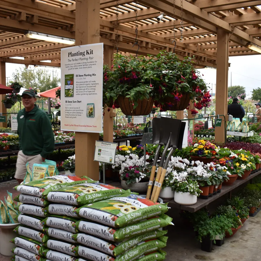 Outdoor garden center with plants, flowers, planting mix bags, and gardening tools under a wooden pergola.