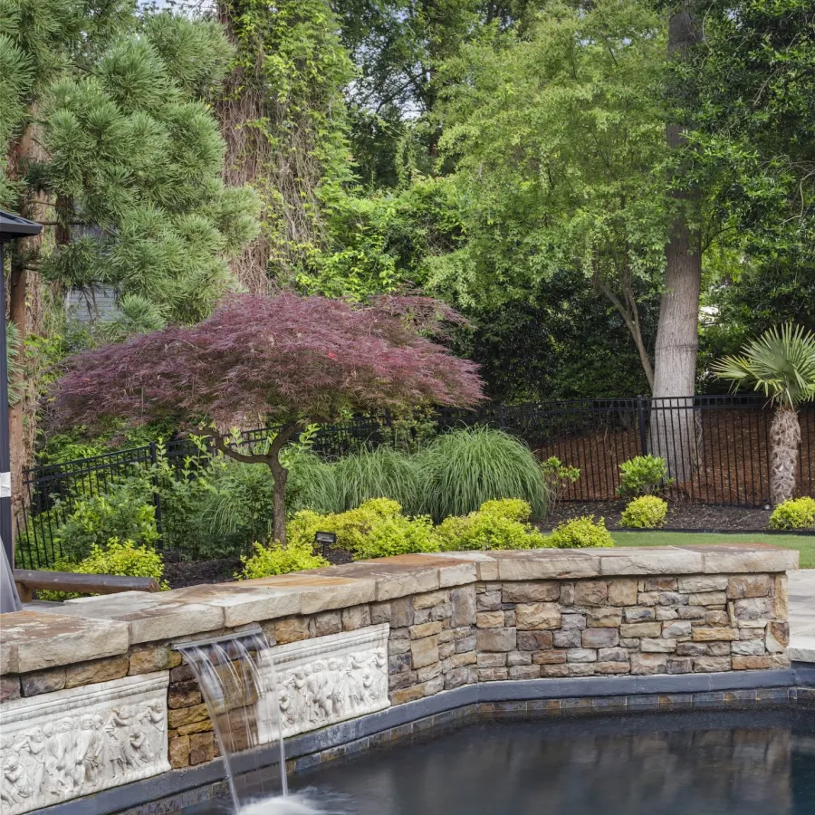 Backyard pool with stone waterfall feature surrounded by lush green trees and colorful landscaping.