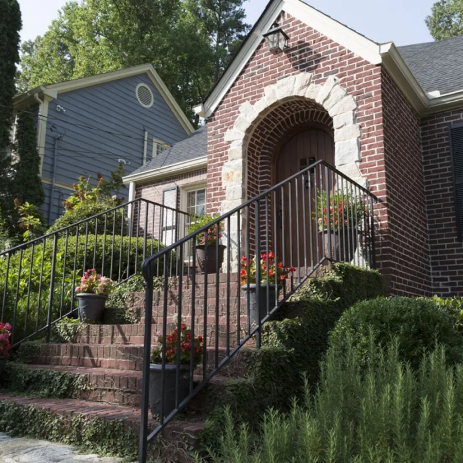 Brick house entrance with stone arch, black railings, flower pots on stairs, and green shrubs on sides