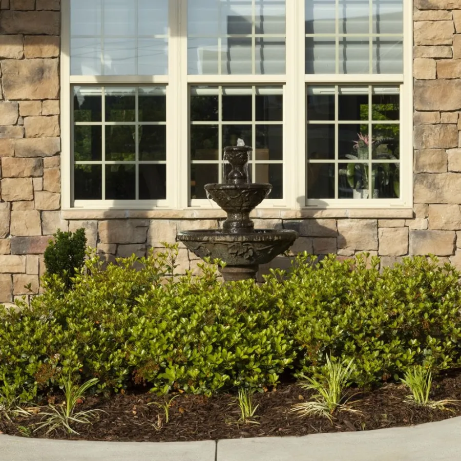 Stone house wall with large windows, central black tiered fountain, green shrubbery, and flower pots on stone pillars.