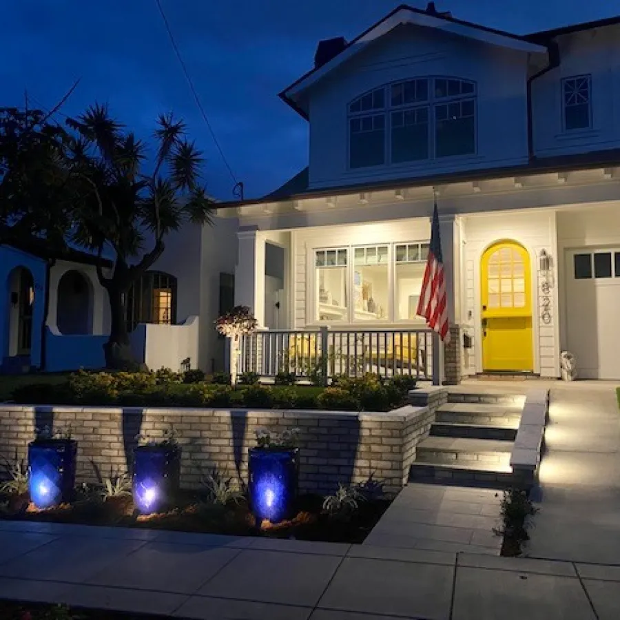 Two-story white house with a bright yellow door, American flag, well-lit porch, and blue garden lights at night.