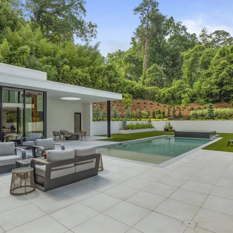 Modern patio with outdoor seating and a pool surrounded by greenery and trees under a clear sky.