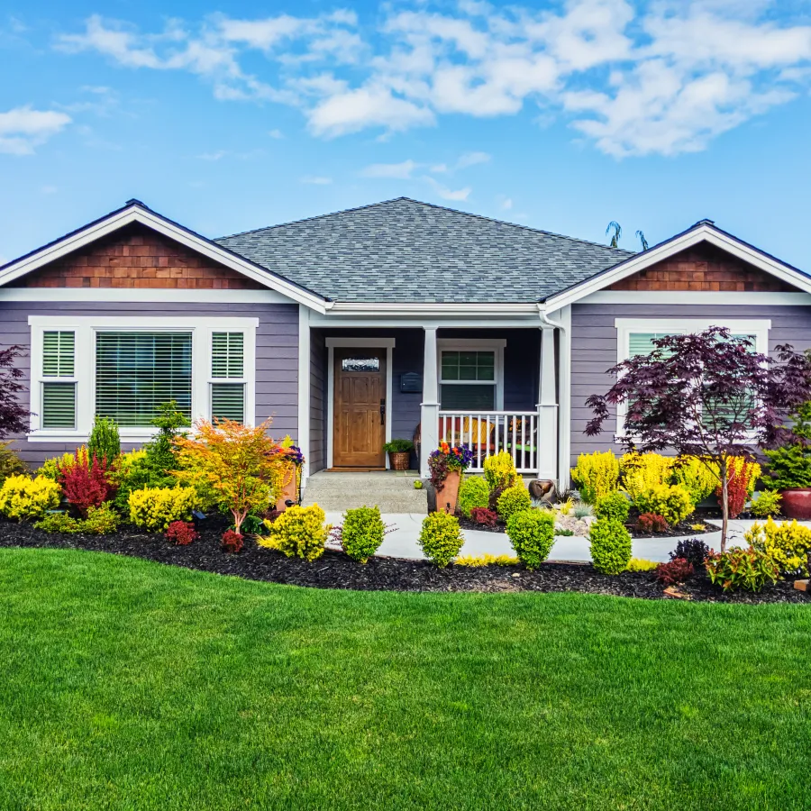 Single-story gray house with a well-maintained colorful garden and lush green lawn under a blue sky.