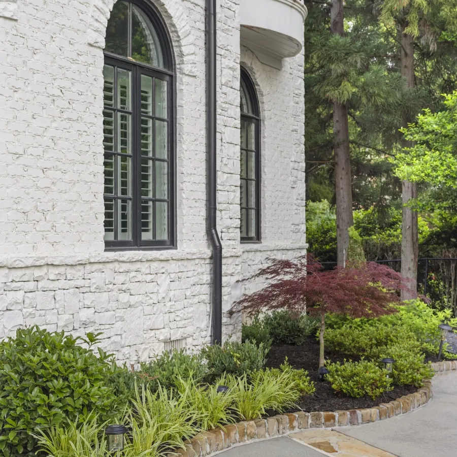 Curved garden bed with lush plants and trees along white brick house with arched windows and stone pathway.