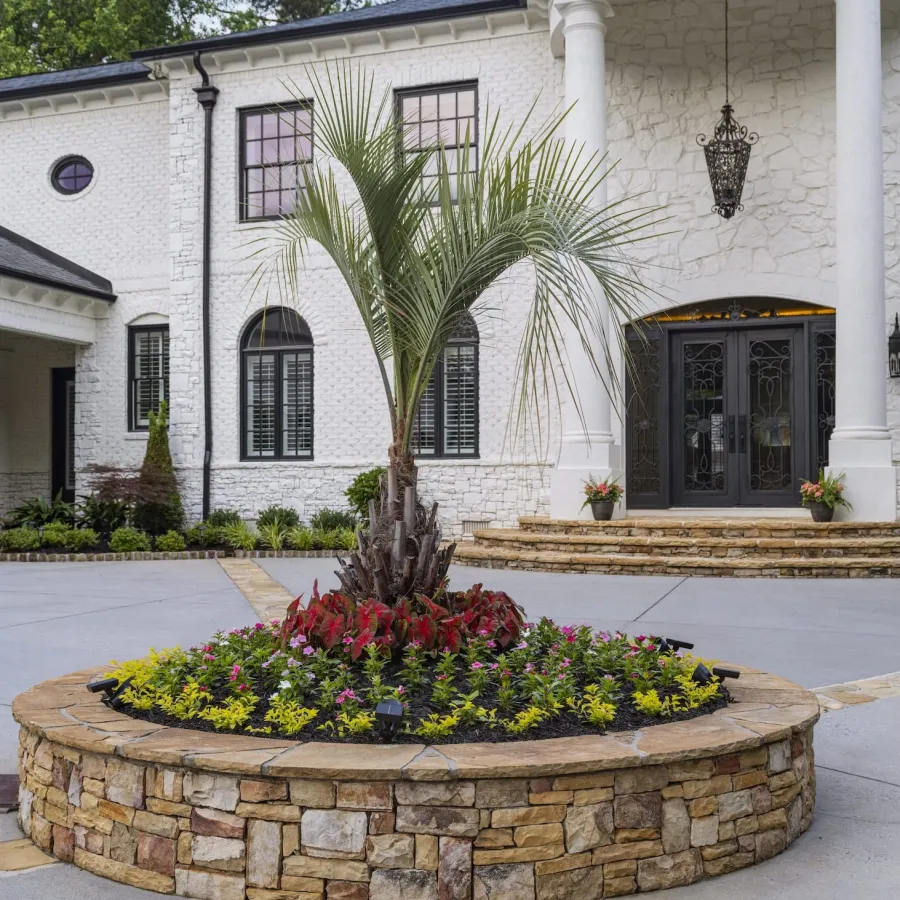 Stone-bordered circular garden bed with palm tree and flowers in front of white house with columns and black doors.