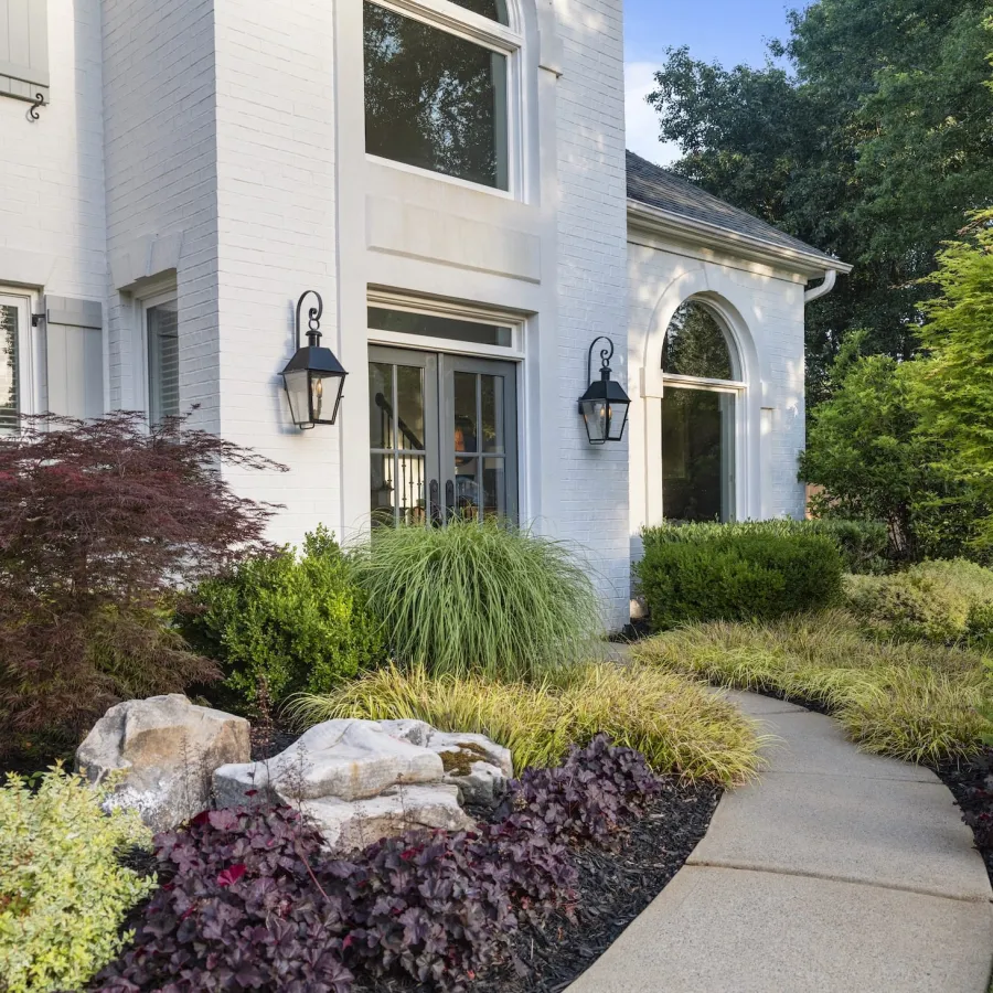 Curved garden path leads to white house entrance surrounded by diverse green shrubs, plants, and decorative rocks.
