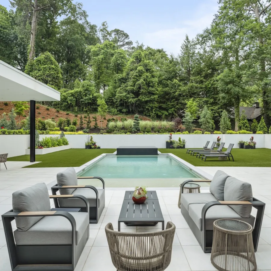 Modern outdoor patio set with grey cushions by a rectangular pool surrounded by green lawn and trees.