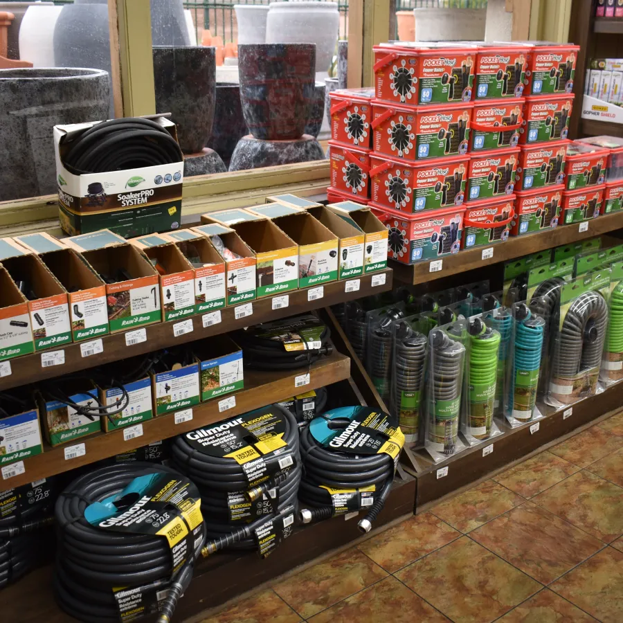 Store shelves stocked with garden hoses, hose connectors, sprinkler systems, and large plant pots in a garden supply shop.