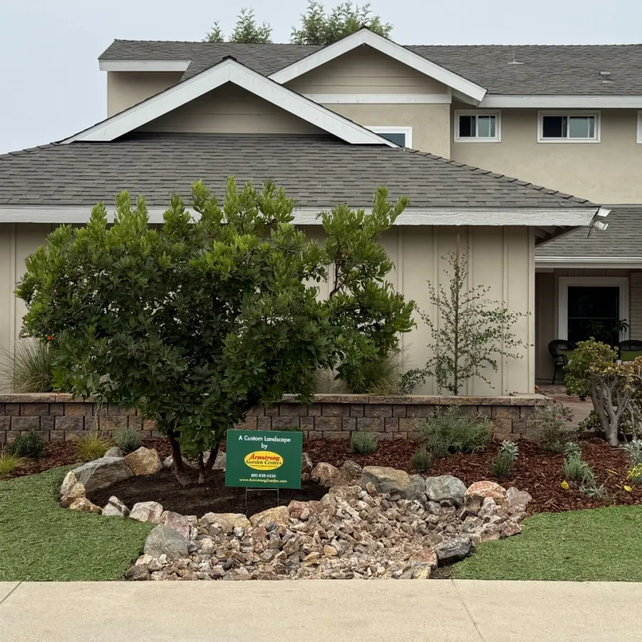 Suburban house front yard with landscaped rocks, green shrub, mulch, and a custom landscaping sign.