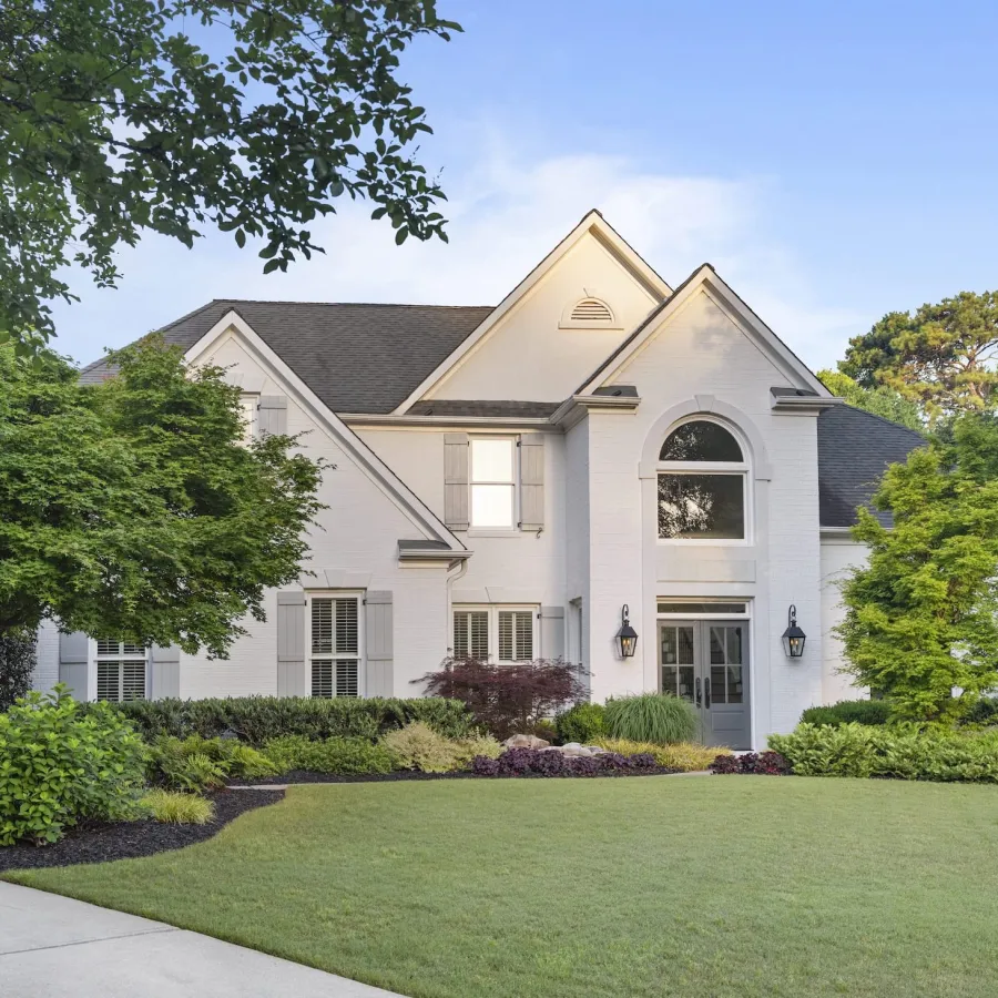 White two-story house with landscaped front yard, green lawn, and mature trees under a clear sky.