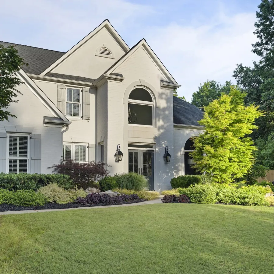 Elegant white two-story home with manicured lawn, shrubs, and bright green tree in front yard on a sunny day